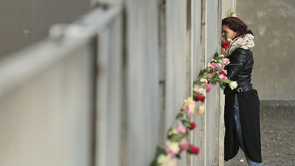 امرأة تقف بجانب جزء من سور برلين وقد وضعت به الورود