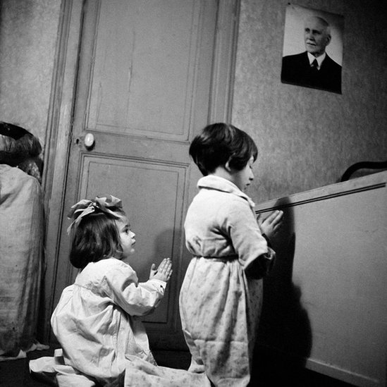 Imagen de dos niñas rezando en una habitación junto a una fotografía de Pétain.