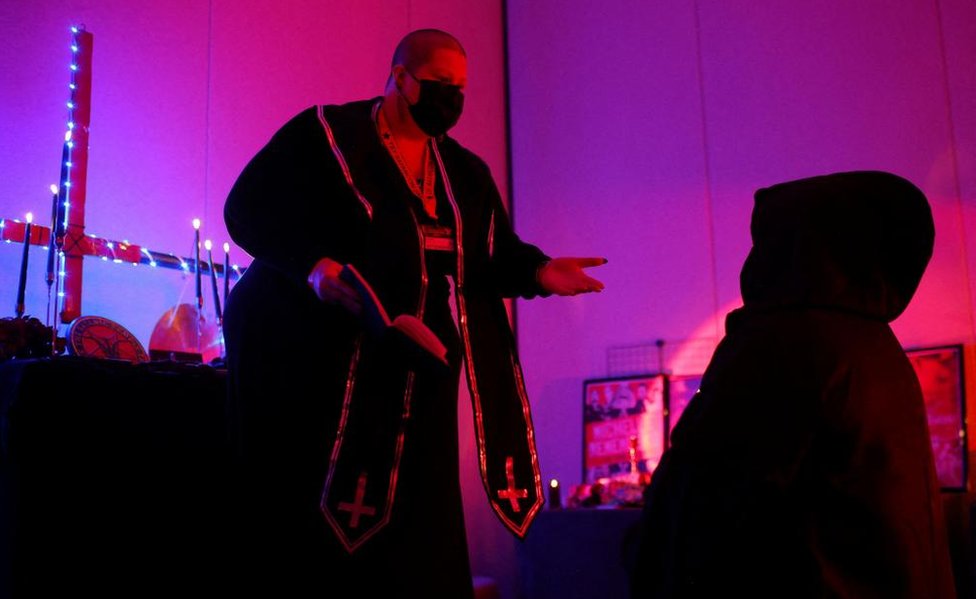 Minister Rose d'Arc performs an Unbaptism Ceremony in a black robe edged with upside down crosses. An altar decked in blue-white lights also with an upside down cross on it stands behind. The subject of the ceremony kneels down in a black robe and hood.