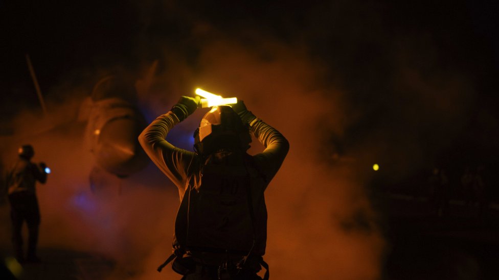 A military jet emerges through condensation while a soldier holds glowsticks for the pilot to be directed, on the deck of USS Dwight D Eisenhower on 22 January 2024