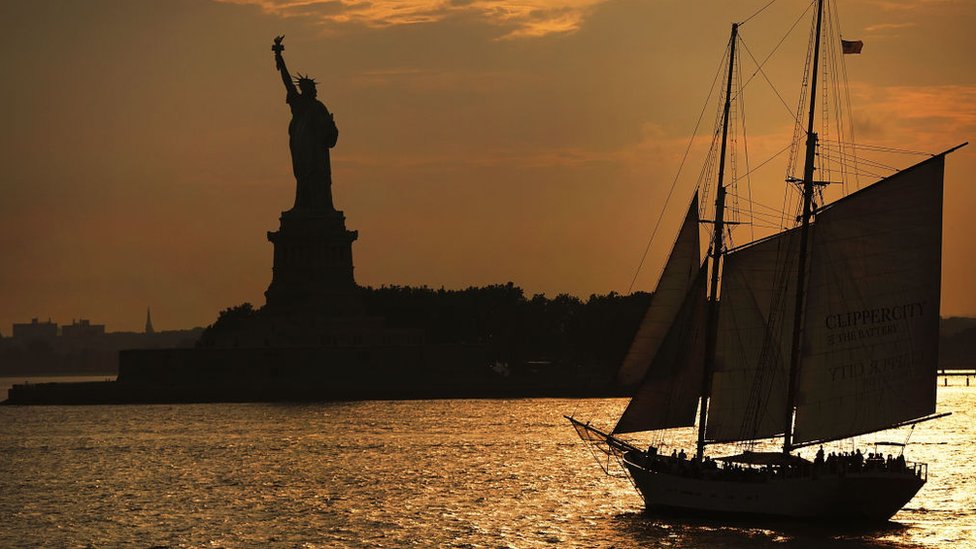 Vista de la estatua de la libertad al atardecer, con un barco antiguo de vela en la bahía