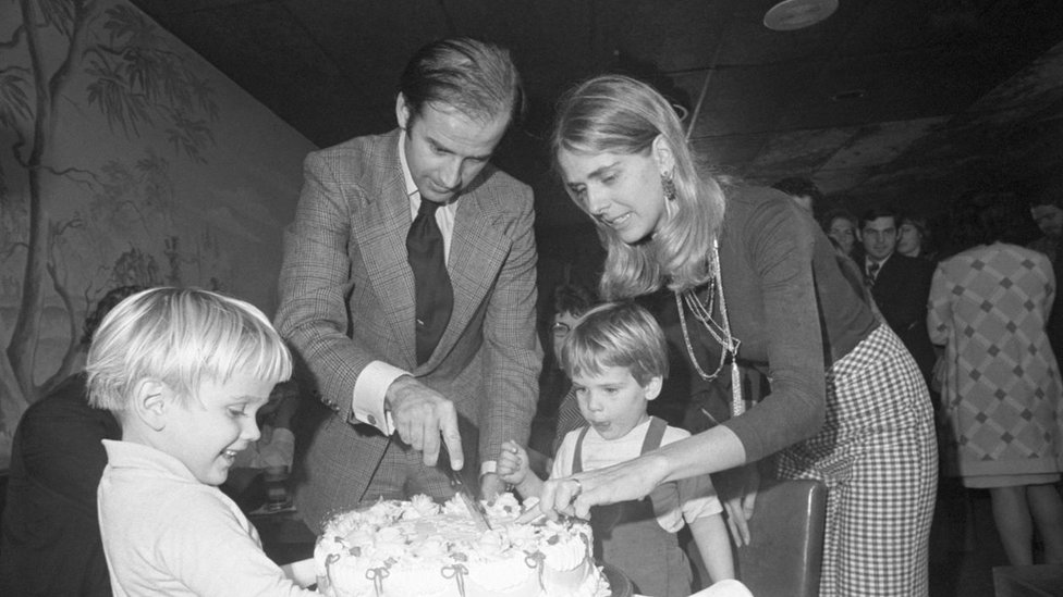 A black and white photo of Joe Biden alongside his first wife and children. They are cutting a cake