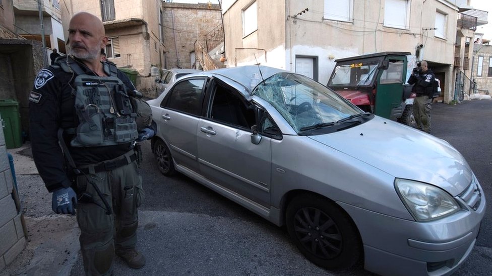 A member of the Israeli security forces stands next to a car in the northern Israeli village of Fassuta that was damaged by a rocket fired from Lebanon (6 April 2023)