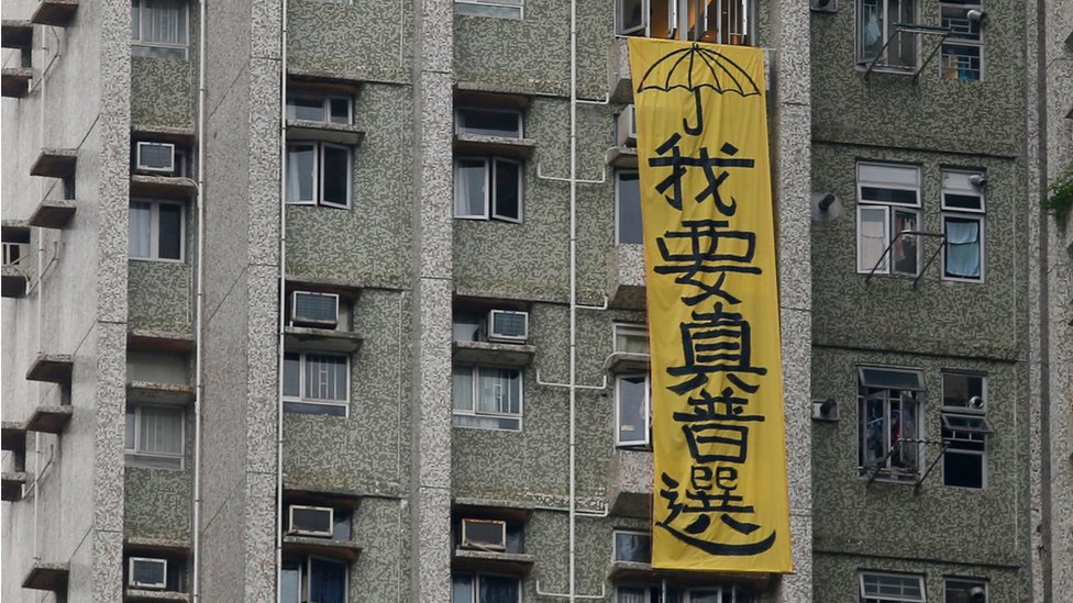 Вертикальный желтый баннер с черным текстом, висящий сбоку от многоквартирного дома, с надписью «Мне нужно настоящее всеобщее избирательное право», в Гонконге, 19 мая