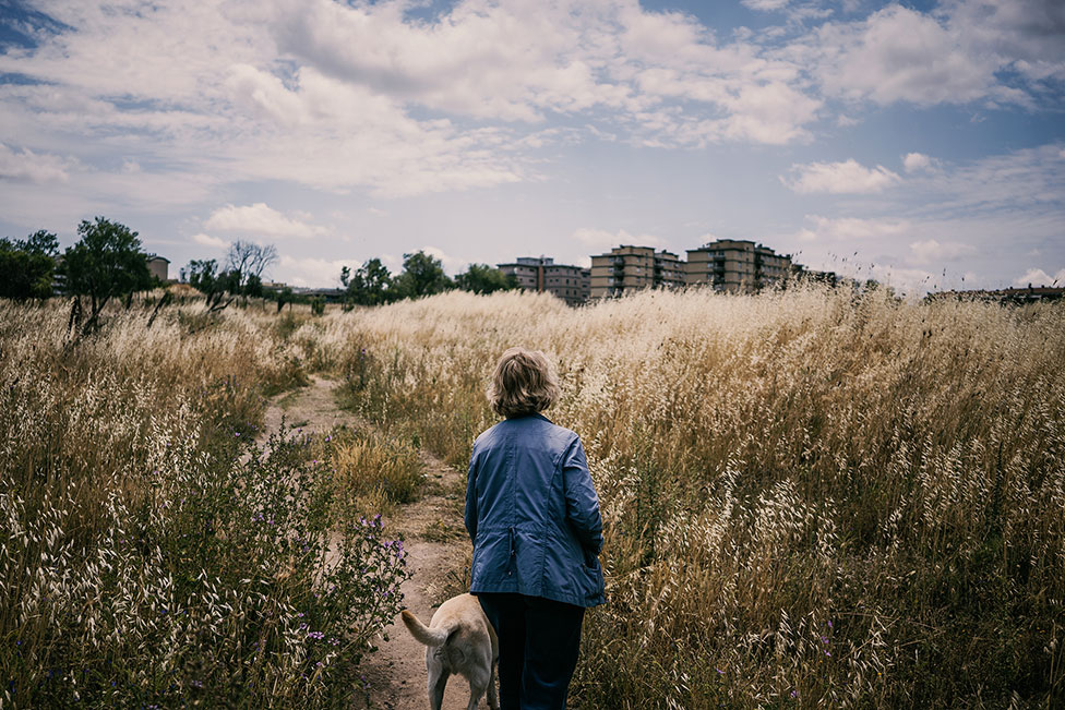 Дама стоит в поле с собакой на поводке рядом с жилым домом