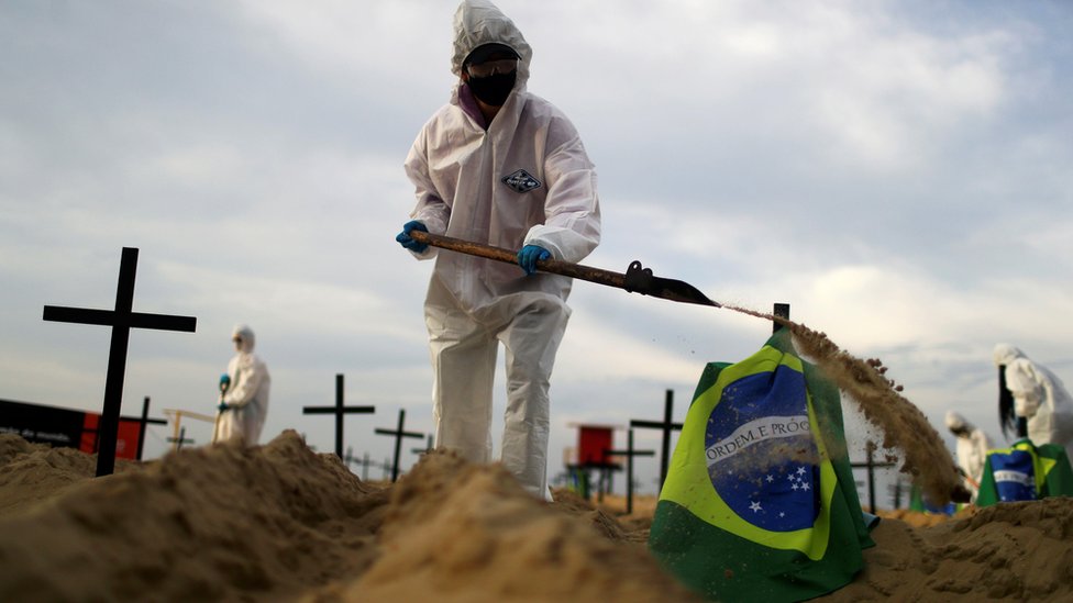 Homem com equipamentos de proteção e uma pá enterrando uma bandeira do Brasil em uma praia do Rio de Janeiro
