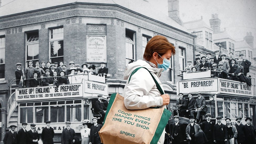 A woman wearing a protective face mask in Chelmsford, England, walks past a large picture showing a demonstration by the National Service league taken sometime before the First World War.