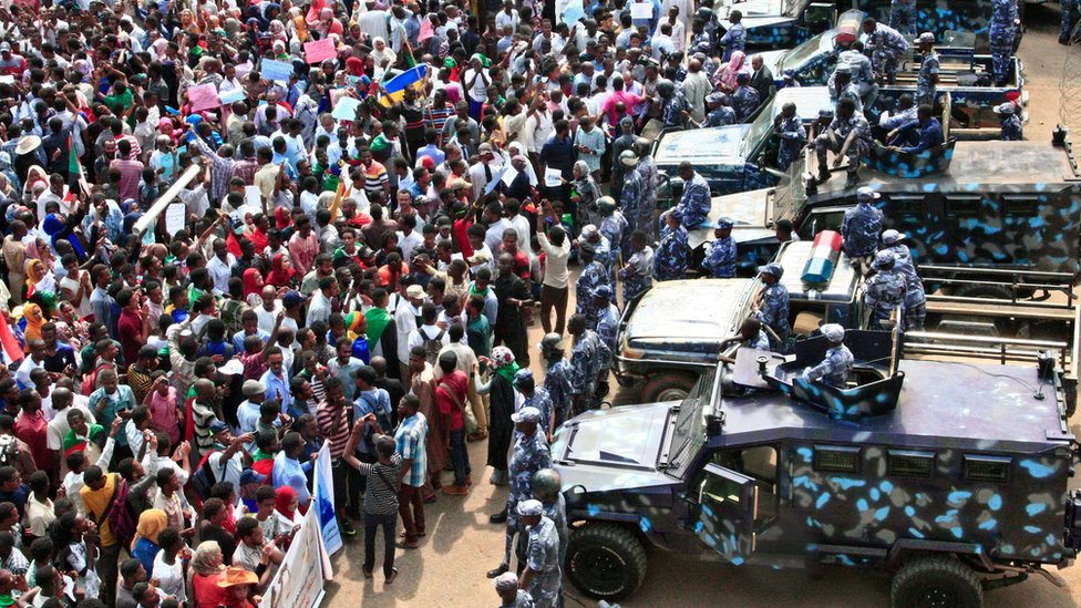 Люди митингуют перед машинами сил безопасности во время массовой демонстрации у президентского дворца в столице Судана Хартуме
