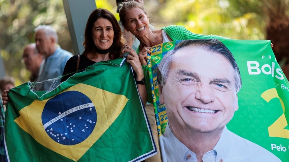 Duas mulheres sorrindo e mostrando para a câmera bandeira do Brasil e imagem de Bolsonaro