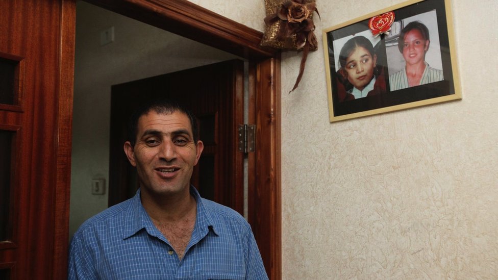 Bassam Aramin standing in a doorway with photos of his daughter and Ram's daughter hanging on his wall