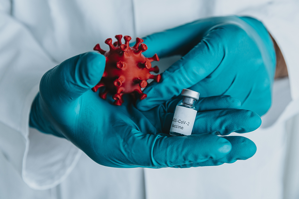 Una persona con guantes sostiene una vacuna y un modelo de un coronavirus