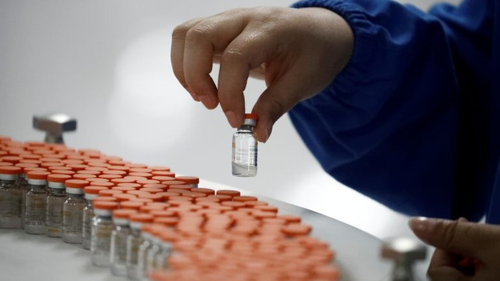 Linha de montagem com vários frascos de vacina, um deles coletado por uma mão