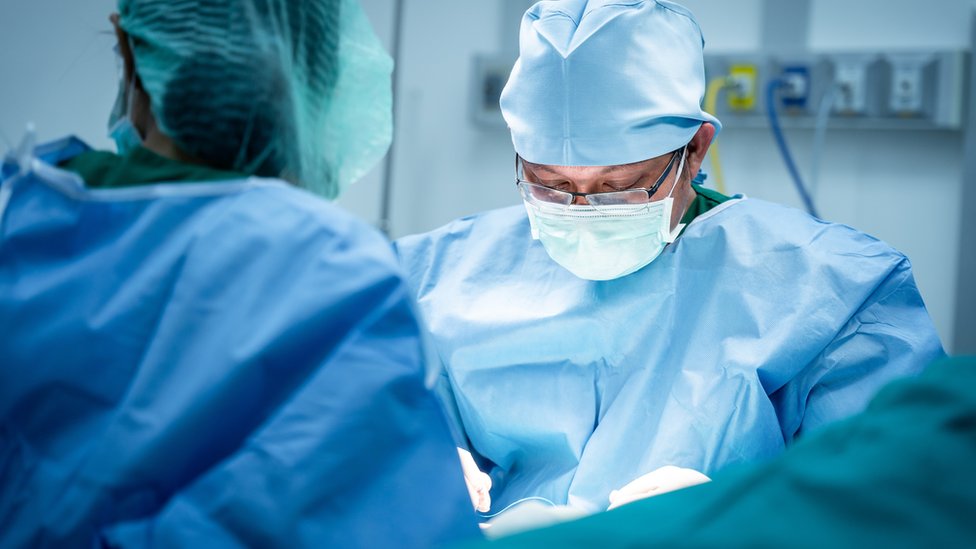 Um cirurgião usando óculos e uma máscara facial é visto olhando para baixo durante uma operação