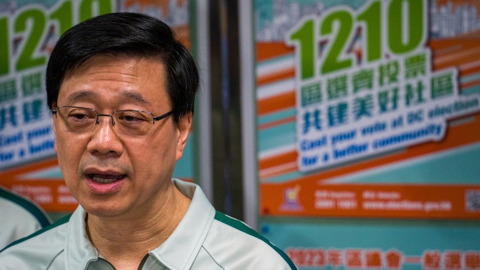 中國香港特首李家超巡視一處公共屋邨之後在區議會選舉宣傳海報前向媒體記者講話（15/11/2023）