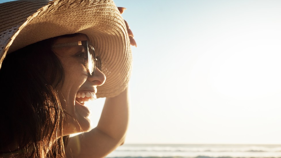 Mujer sonriendo en la playa con sombrero de ala ancha y lentes de sol
