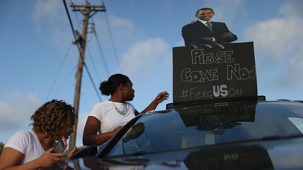 Демонстранты несут табличку с изображением Барака Обамы, читающего «Пожалуйста, приходите сейчас» 17 августа 2014 года в Фергюсоне