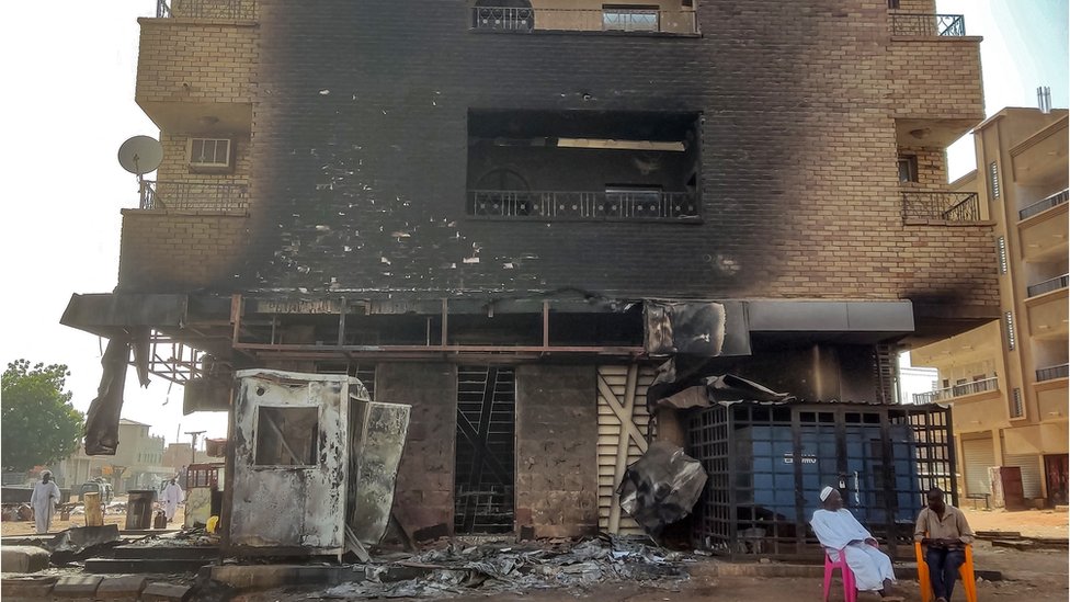 Building charred from an attack with the ground floor completely destroyed. Two men sit on colourful bright chairs at the bottom of the building.