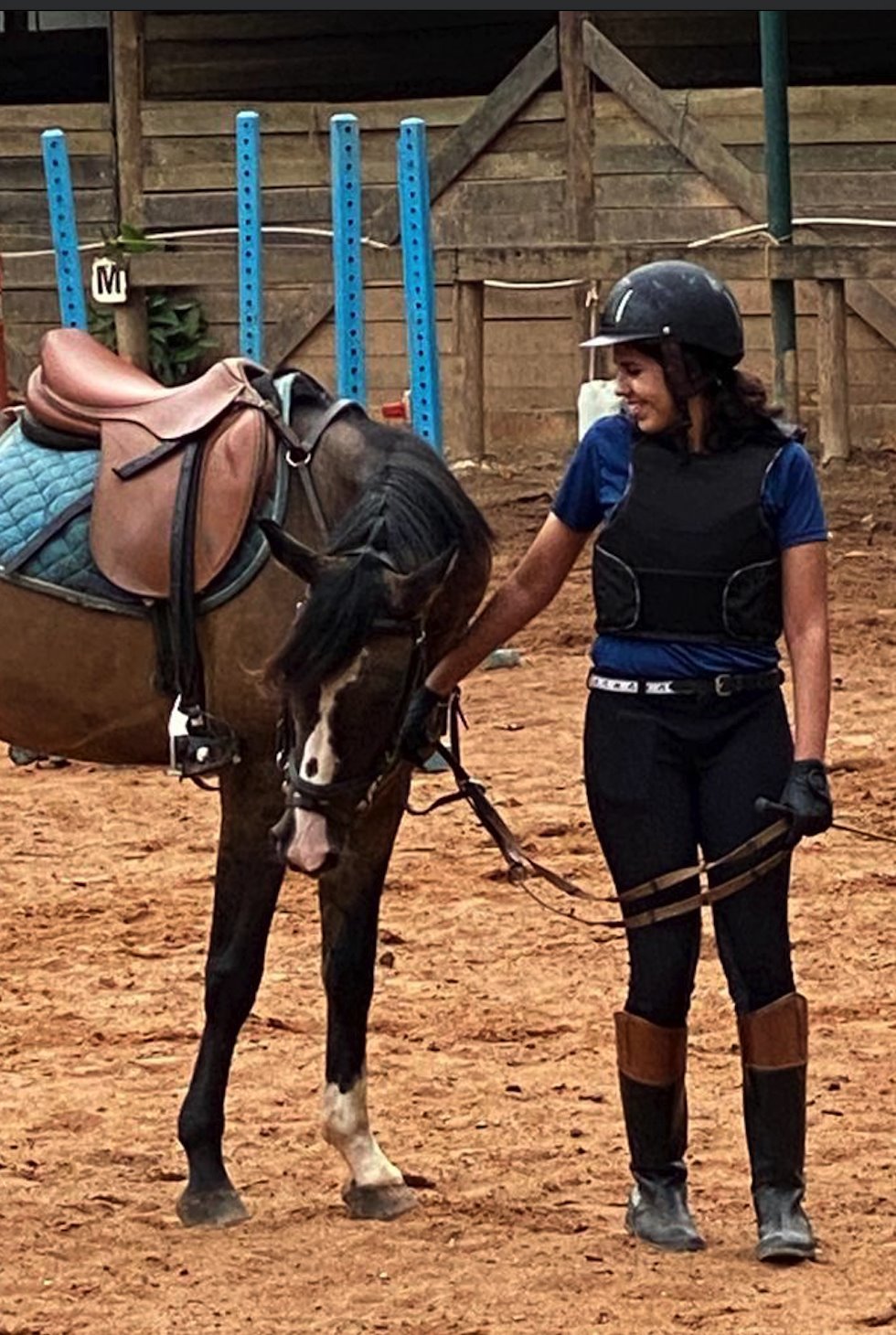 Larissa em pé em um chão de areia de um haras, enquanto brinca com um cavalo