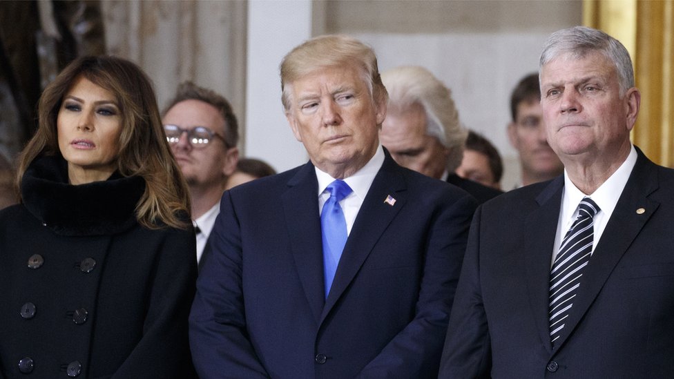Президент Дональд Трамп и первая леди Мелания Трамп встают с Франклином Грэмом во время церемонии