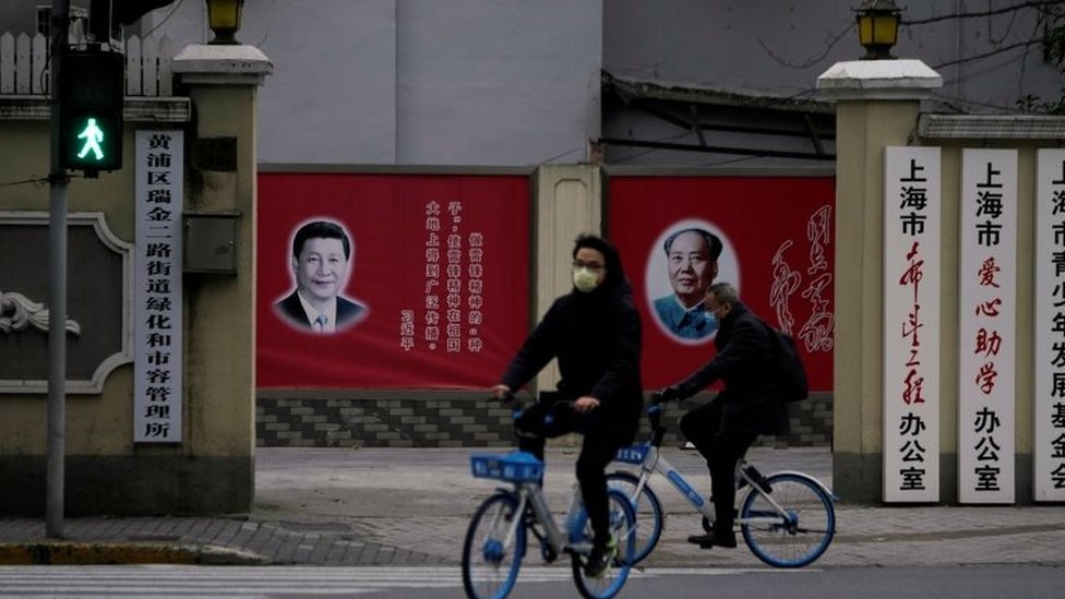 أشخاص يرتدون أقنعة يمرون من امام صور للرئيس الصيني شي جين بينغ والرئيس الصيني الراحل ماو تسي تونغ