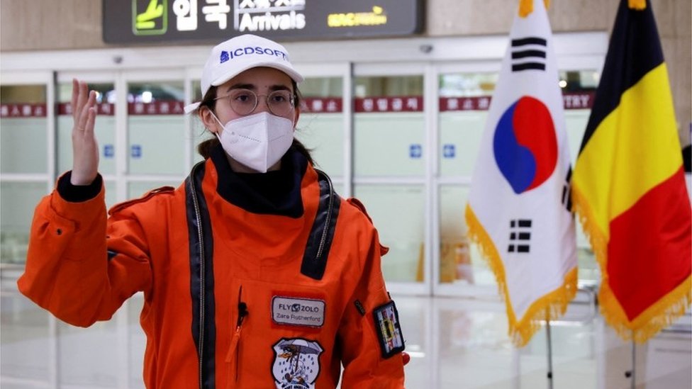 Zara Rutherford de pie en la sala de llegadas del Aeropuerto Internacional de Gimpo en Seúl con un traje de inmersión naranja y una máscara blanca mientras es entrevistada por periodistas.