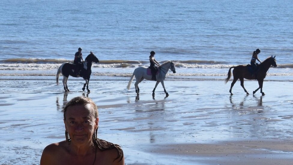 Donna Price nua na praia com três pessoas cavalgando atrás dela