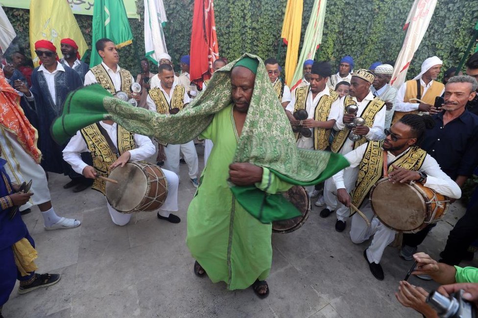 رجل يرتدي الزي الأخضر التقليدي يرقص وسط دائرة من الرجال الذين يرتدون الملابس البيضاء والصفراء التقليدية ويعزفون على الطبول