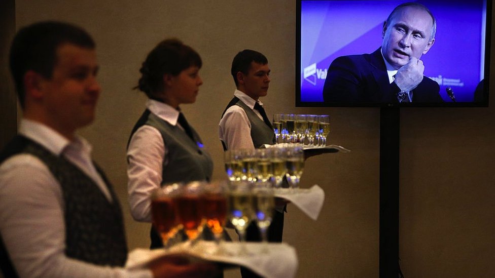 Waiters hold trays of drinks while Putin appears on a TV screen