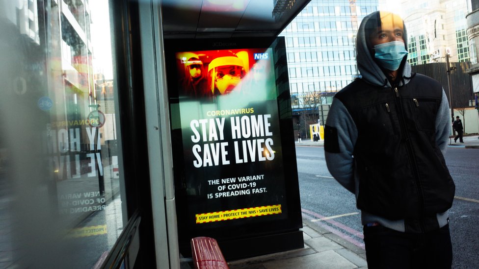 A man wears a mask at a bus stop with a Stay Home Save Lives poster