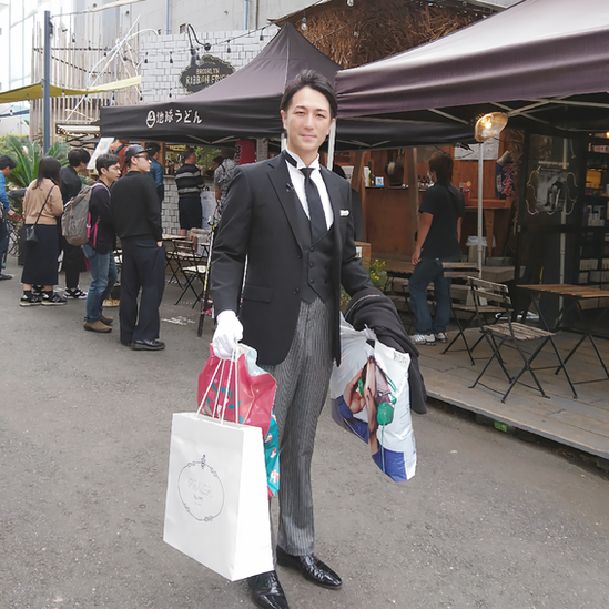 Yuichi Ishii vestido elegantemente de saco y chaleco negros, guantes blancos, cargando bolsas de compras