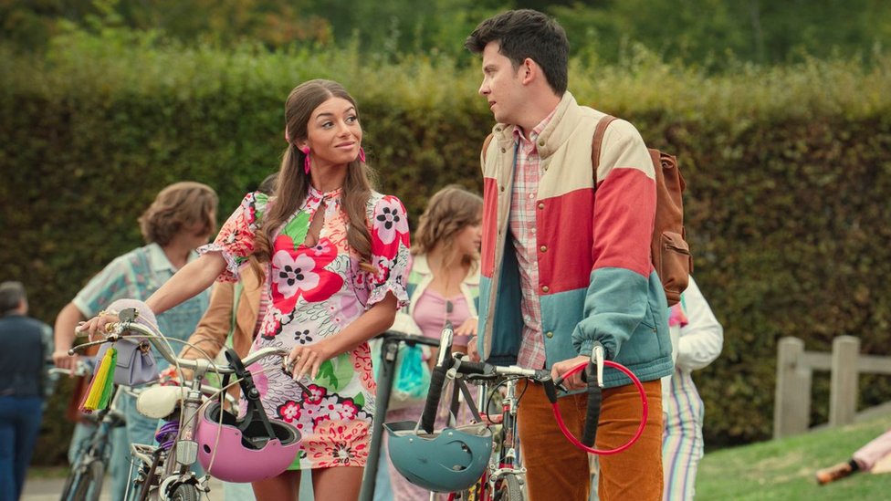 Mimi Keene as Ruby and Asa Butterfield as Otis in Sex Education. Ruby and Otis are speaking to each other outside as they wheel their bikes. Ruby, a young woman with long light brown hair, wears a short floral dress and large pink earrings. Otis, a young man with short dark hair, wears orange cord trousers, a pink plaid shirt and a blue, cream and red striped puffer jacket.