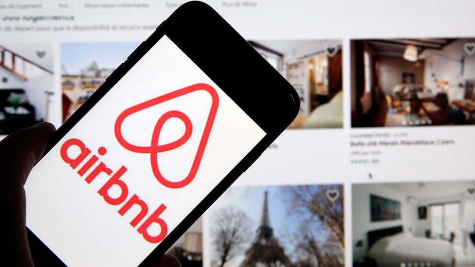 Мужчина держит телефон с логотипом Airbnb перед веб-сайтом компании