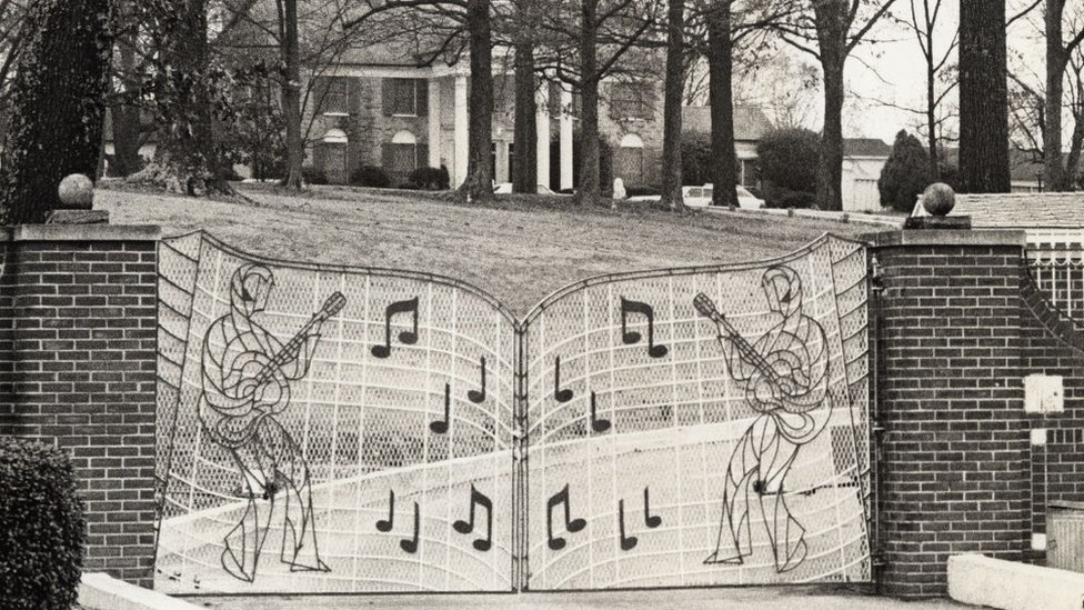 Foto em preto e branco do portão, curvado e com duas figuras de ambos os lados semelhantes ao rei do rock, com um violão, calça flare e peruca e várias notas musicais grandes ao redor