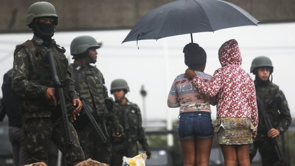 Молодые люди стоят рядом с бразильскими солдатами во время «мегаоперации», проводимой Бразильскими вооруженными силами вместе с полицией против членов банд в семи самых жестоких общинах «фавел» Рио, 21 августа 2017 года в Рио-де-Жанейро, Бразилия.