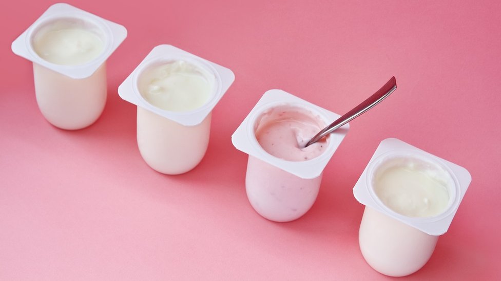 Concept image of better choice: four yogurts in white plastic cups on pink background with copy space. Strawberry pink yoghurt with spoon in it.