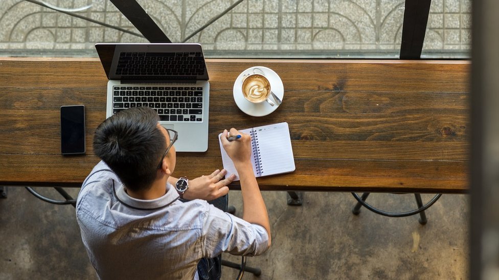 رجل أعمال شاب يدون ملاحظات إلى جانب جهاز كمبيوتر محمول على طاولة خشبية