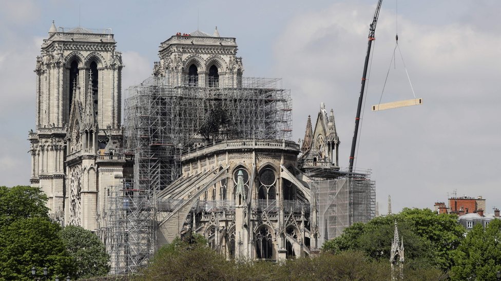 Пожарные работают на вершине башни собора Нотр-Дам, пока кран поднимает строительные материалы в Париже 17 апреля 2019 года,