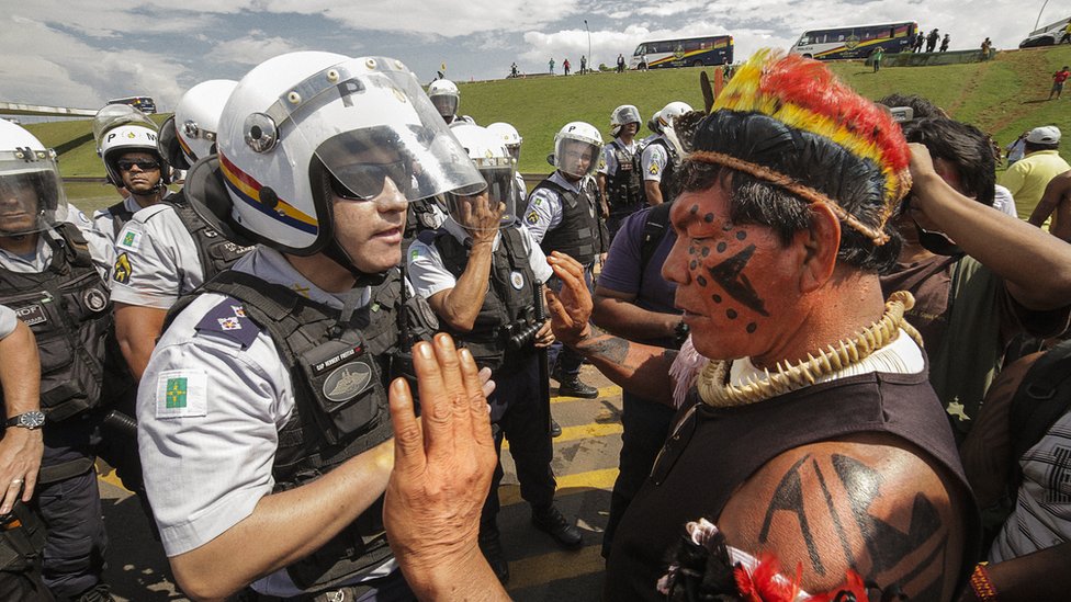 Fotografia colorida mostra grupo de indígenas em frente a policiais paramentados