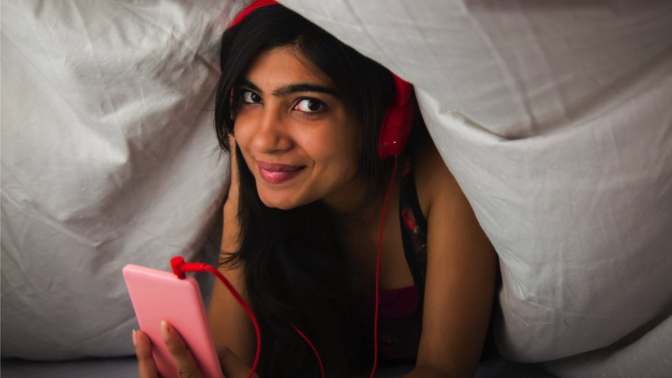 A young woman listening to her phone under a duvet