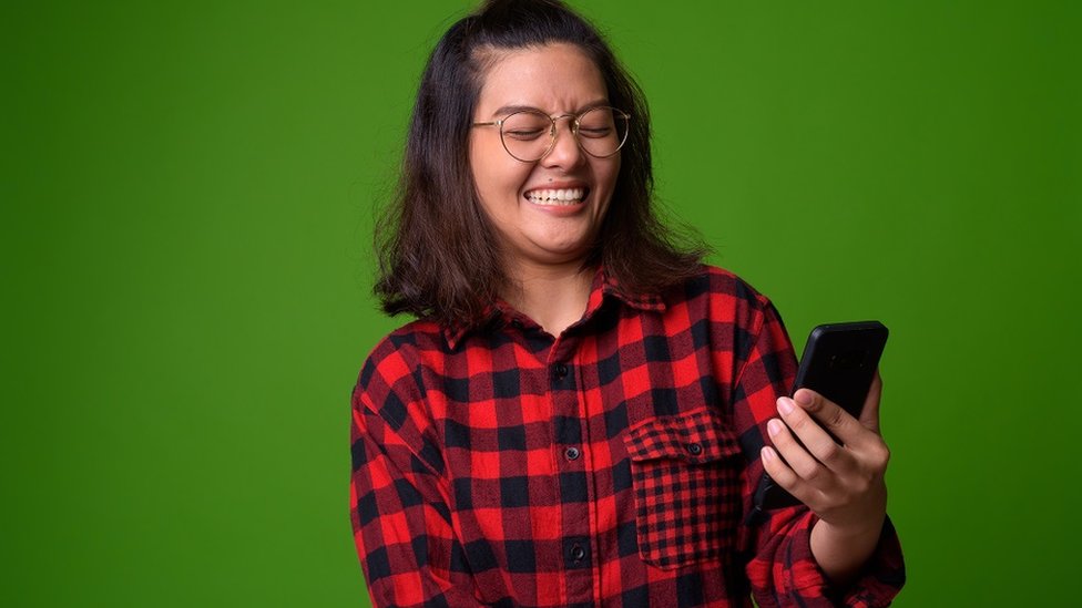 Young woman with a checked red and black shirt. She's laughing and holding a smarphone. Bright green background.