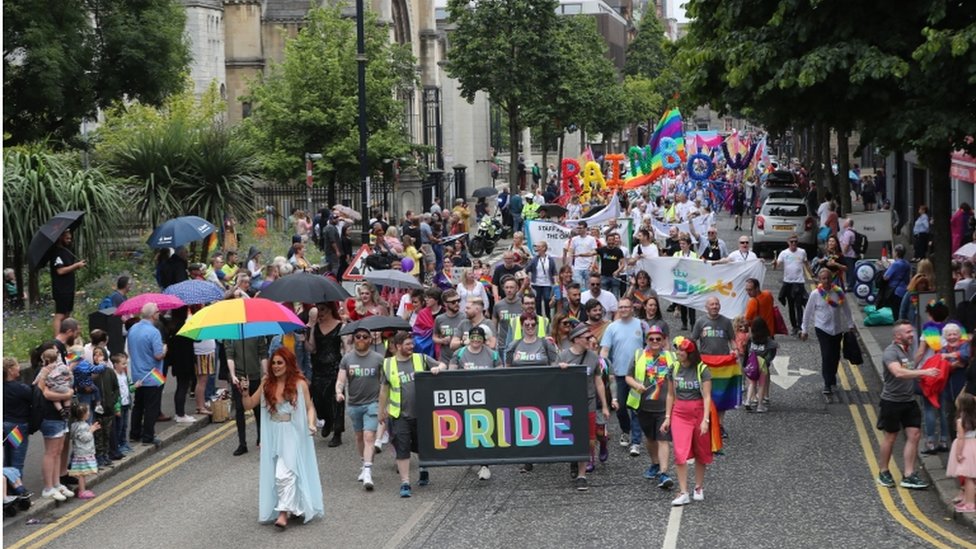 Участники марша держат знамя с надписью BBC Pride, проезжая мимо собора Святой Анны в Белфасте