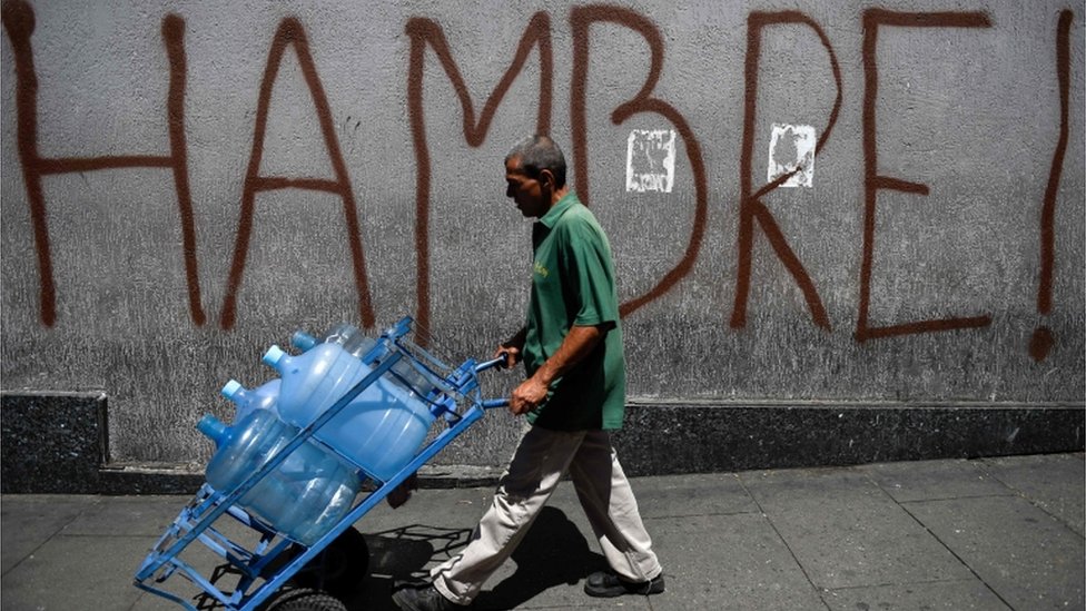 Hombre camina en Venezuela con botellones de agua vacíos frente a una inscripción que dice: Hambre.