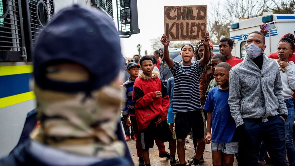 Житель держит плакат с надписью: «Убийца детей» рядом с сотрудниками Южноафриканской полицейской службы (SAPS) возле офисов SAPS в парке Эльдорадо, недалеко от Йоханнесбурга, 27 августа 2020 г., во время акции протеста членов сообщества после 16 Сообщается, что -летний мальчик мертв