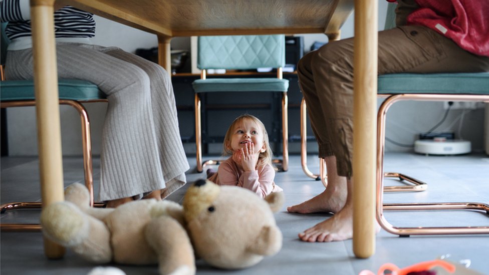 Criança sorrindo debaixo de mesa, onde pode se ver também as pernas de pais e mães sentados