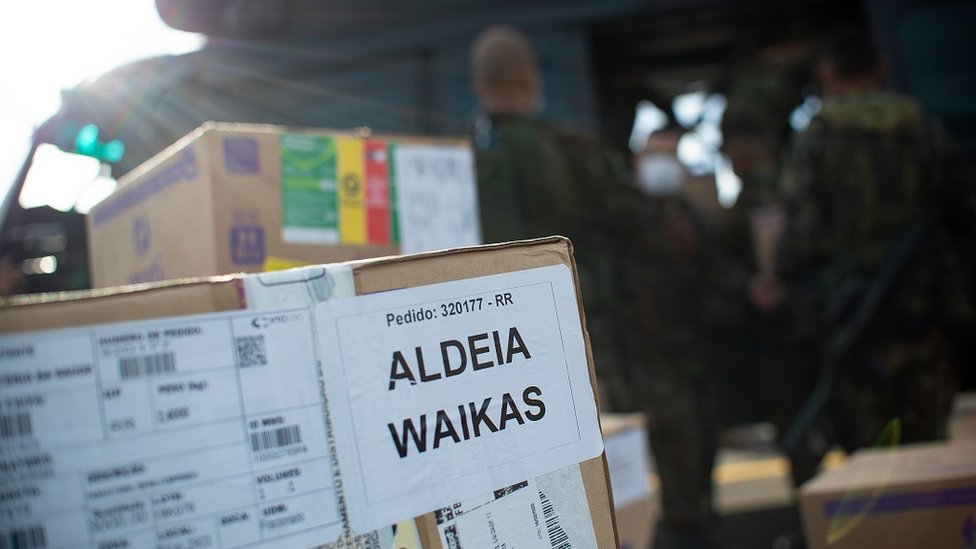 Caixa com aviso 'Aldeia Waikas' em pátio de decolagem, com militares e helicóptero no plano de fundo