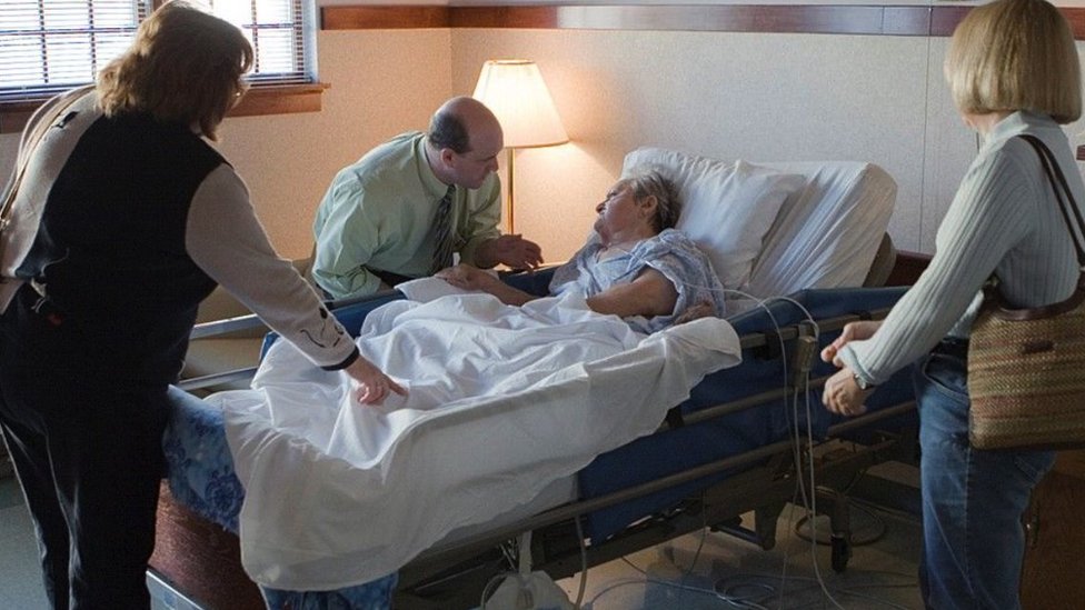 Žena u bolničkom krevetu okružena porodicom