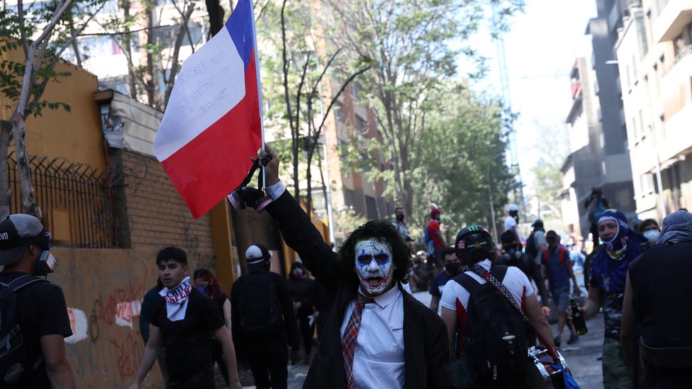 Un hombre con la cara pintada de blanco sosteniendo una bandera de Chile.