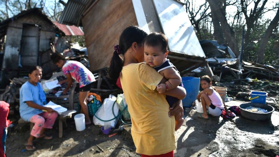Una madre sostiene a su hijo, mientras otros miembros de la familia recogen lo que queda de su casa destruida