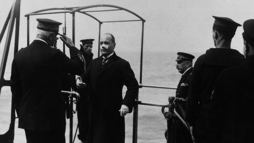 The head of the Fascist government Benito Mussolini welcomed by the admiral while boarding battleship Cavour with a bandaid over his nose.