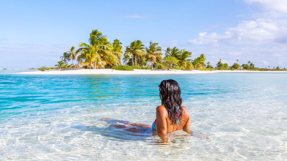 Una mujer de espaldas dentro de un mar cristalino observando una idílica isla en la distancia